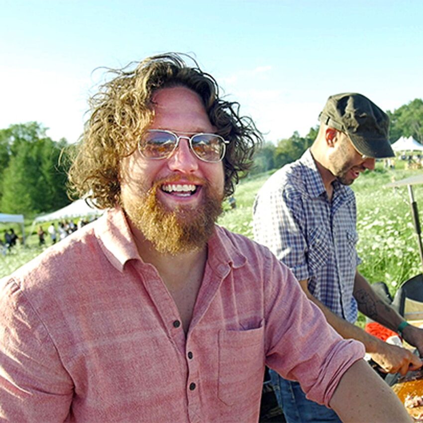 Smiling man with curly hair and glasses outdoors, another person in background with a cap preparing food, grassy field setting.