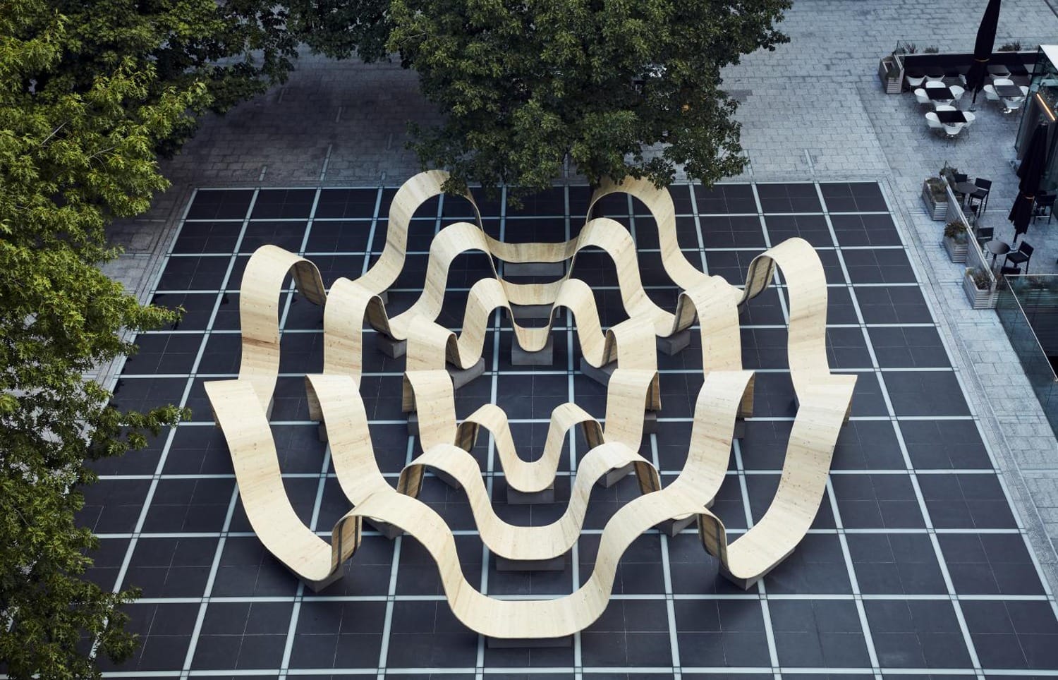 Modern wooden art installation with wavy benches on a black tiled surface surrounded by trees in an urban park setting.