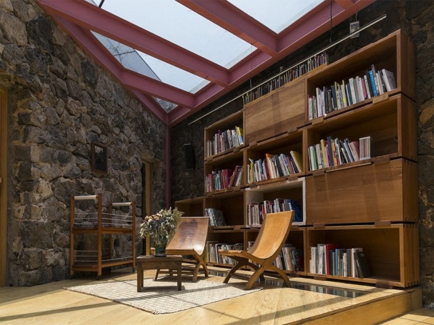 Modern library room with stone walls, wooden shelves full of books, two wooden chairs, a coffee table, and a skylight ceiling.