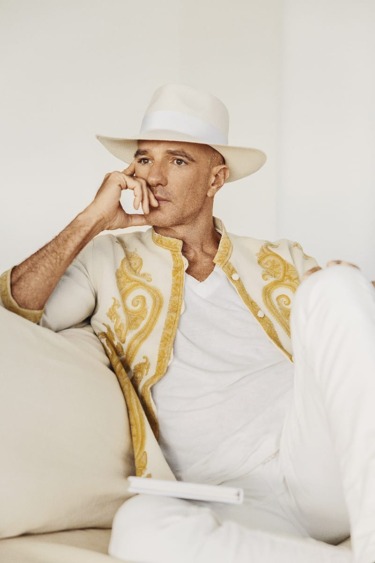 Man in a stylish white and gold jacket with a white hat sitting thoughtfully on a beige couch.