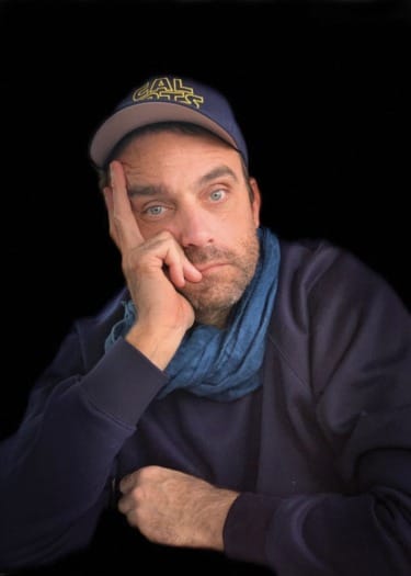 Man wearing a blue cap and scarf, resting his face on his hand against a dark background.