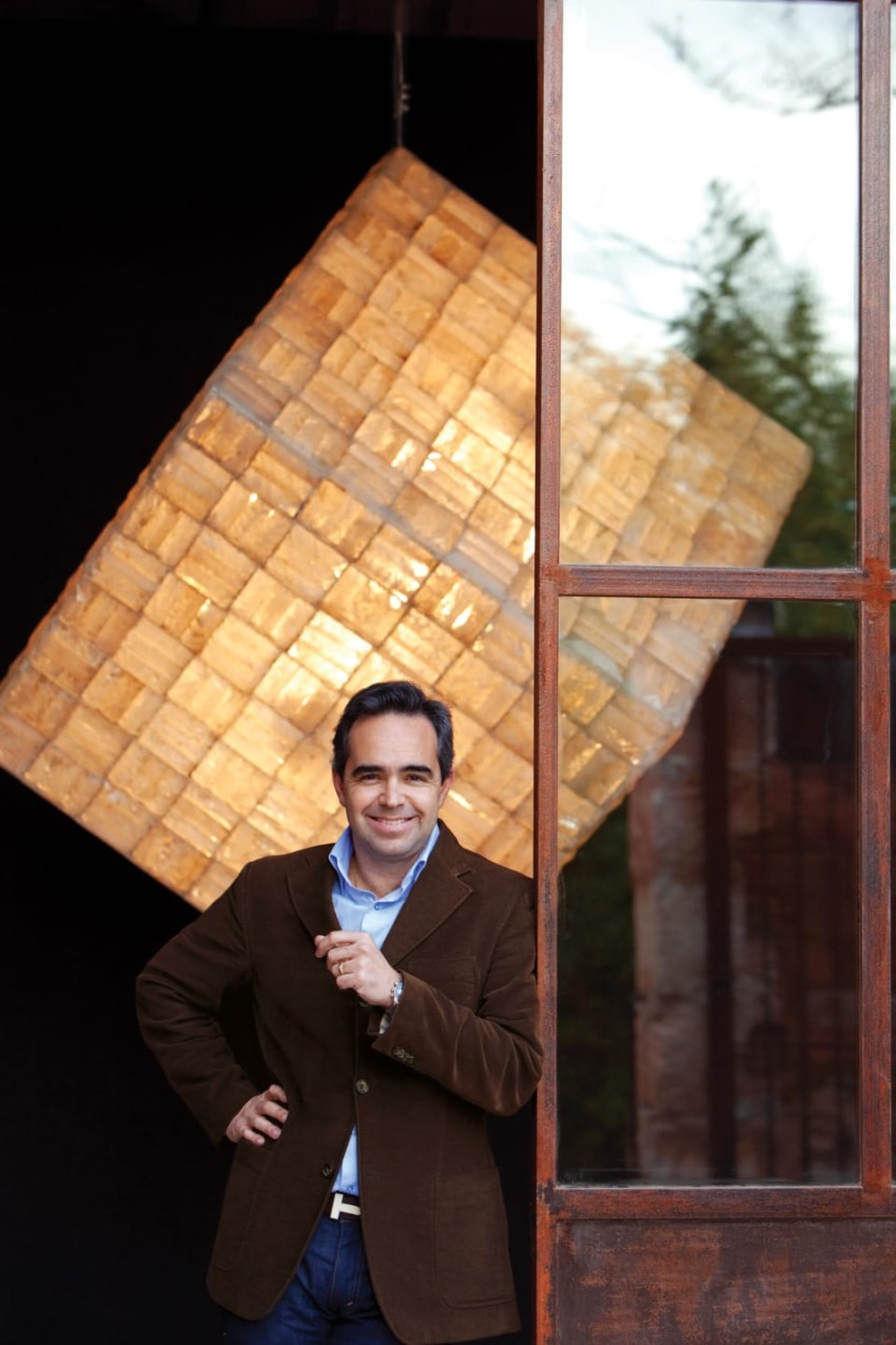 Smiling man in a brown jacket stands by a large cube-shaped light fixture near a rustic wooden door.