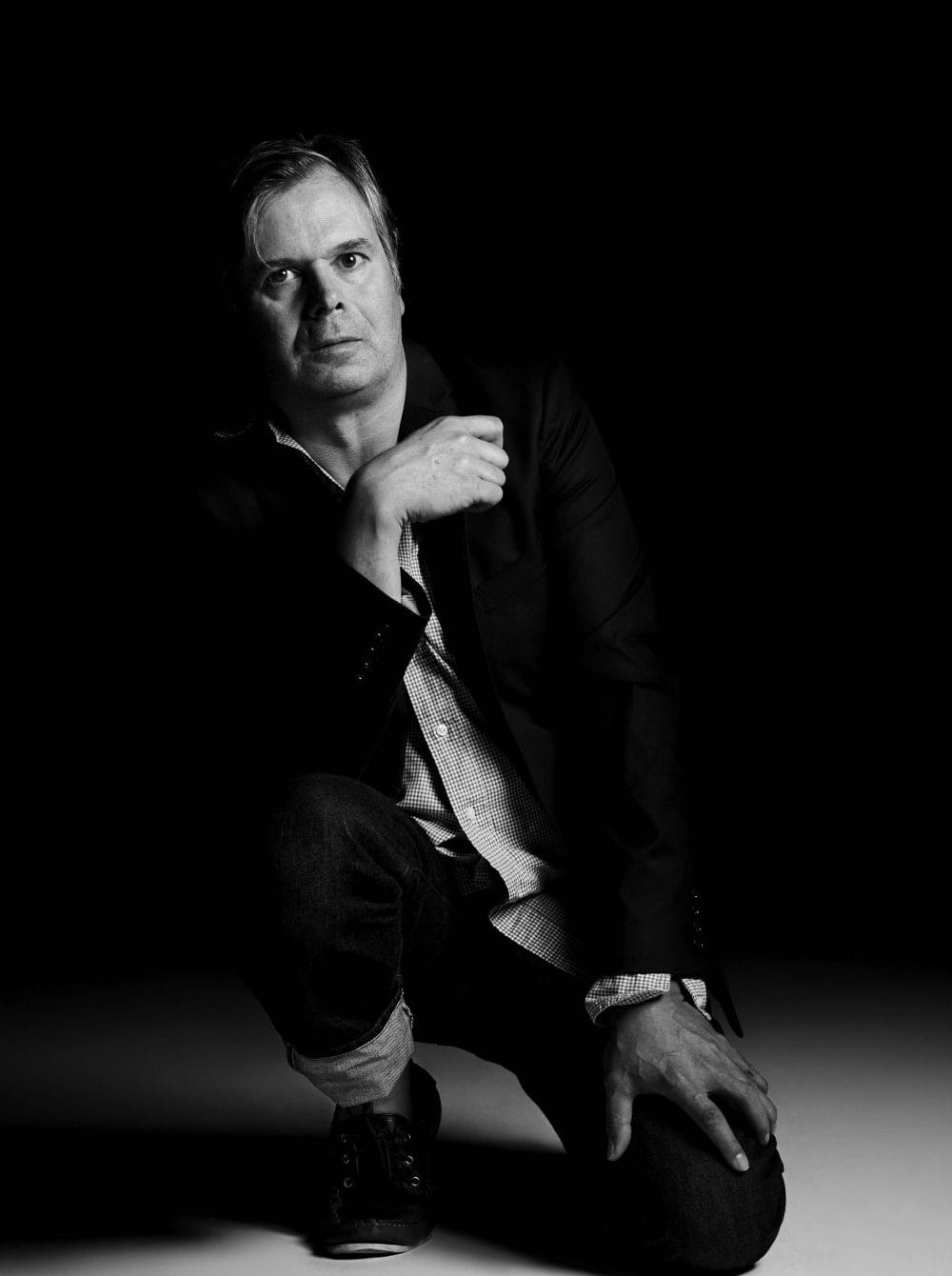 Man in a dark blazer and jeans kneeling on one knee in a dramatic black and white setting.