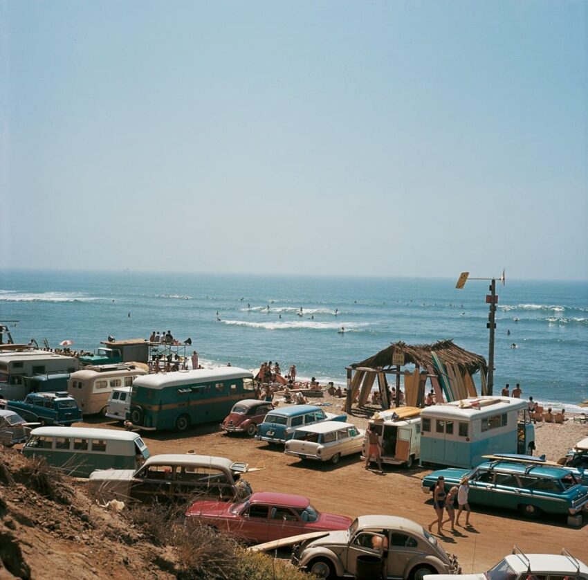 Vintage beach scene with classic cars parked near the shore, surfers and beachgoers enjoying a sunny day by the ocean.