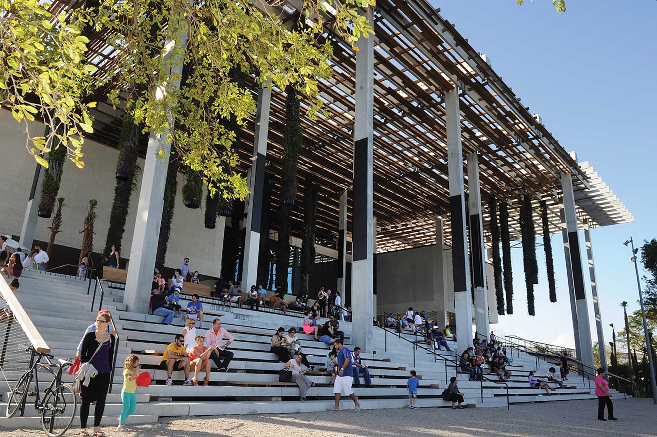 People sitting on outdoor steps under modern architectural canopy with sunlight filtering through, surrounded by trees.