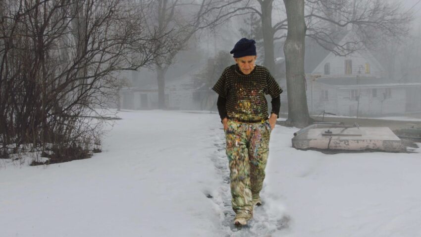 An elderly man wearing paint-splattered clothes walks along a snowy path with trees and mist in the background.