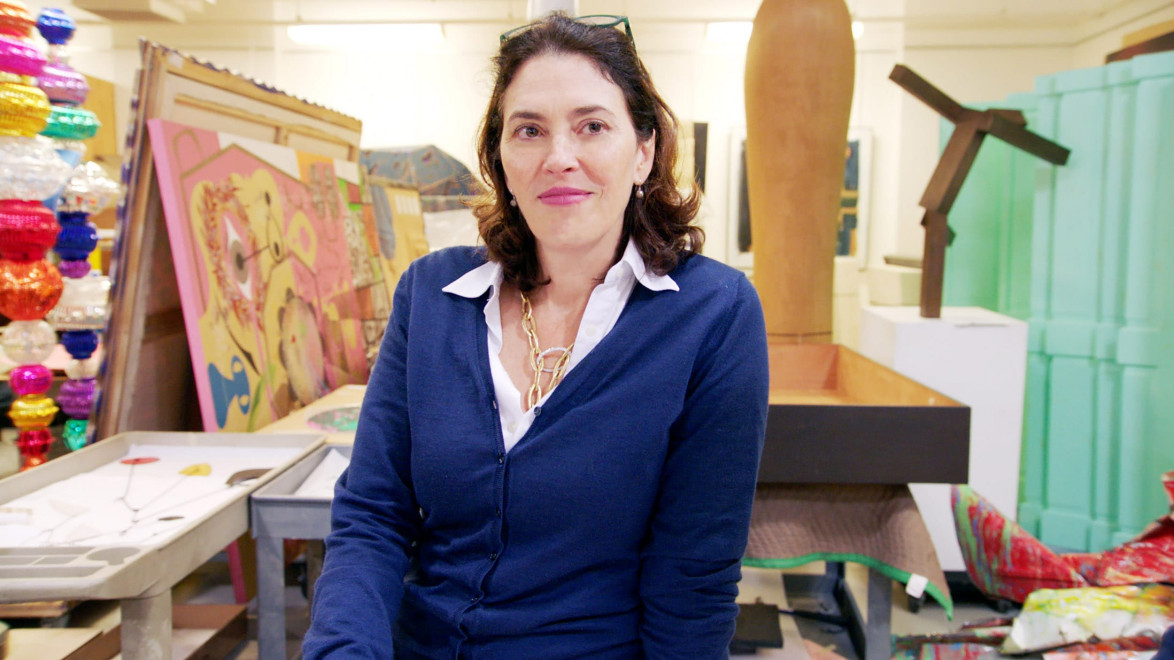 Person in a blue sweater sitting in an art studio with colorful paintings and sculptures in the background.