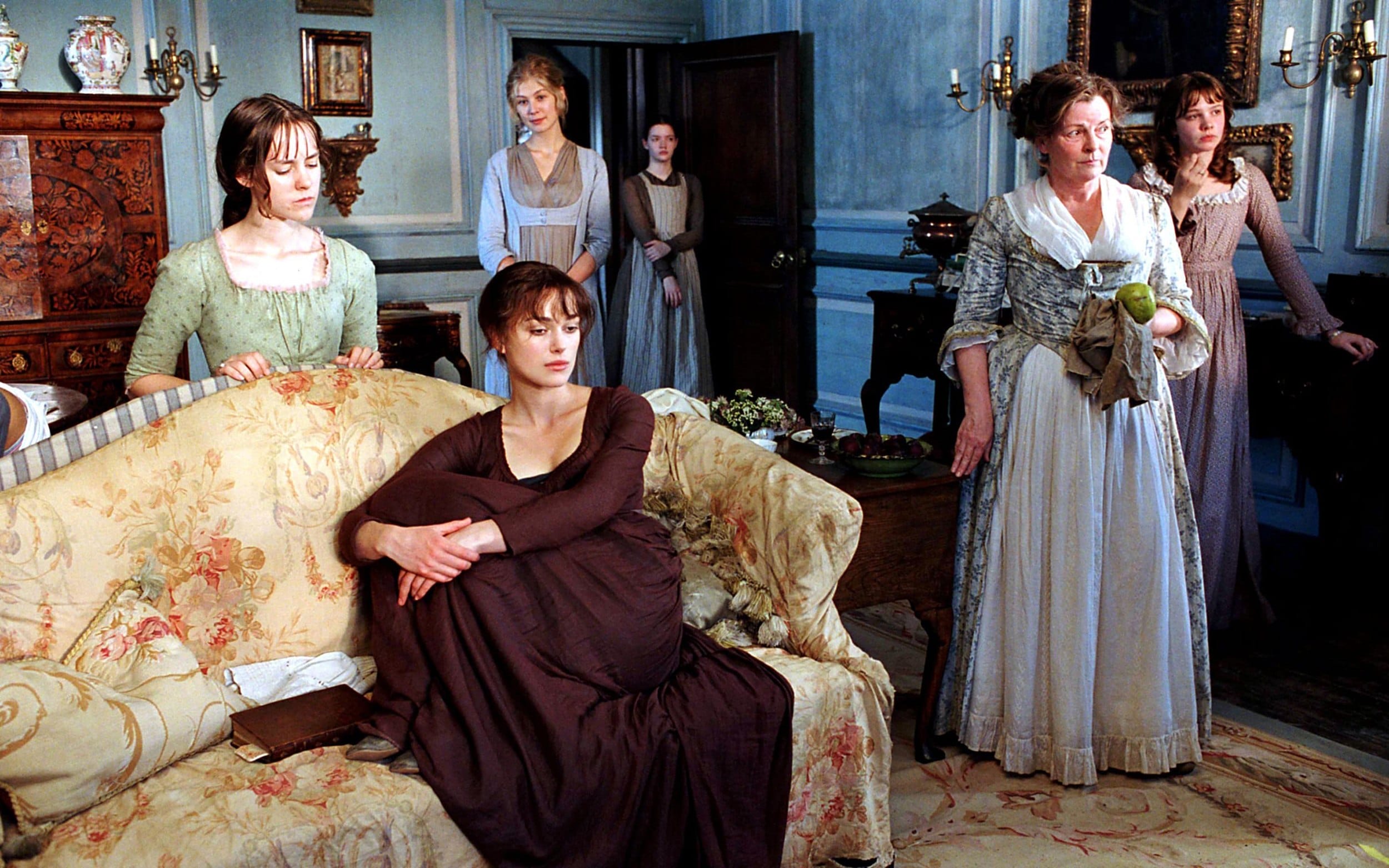 Group of women in period costumes standing and sitting in a vintage-style room, with one woman holding an apple.