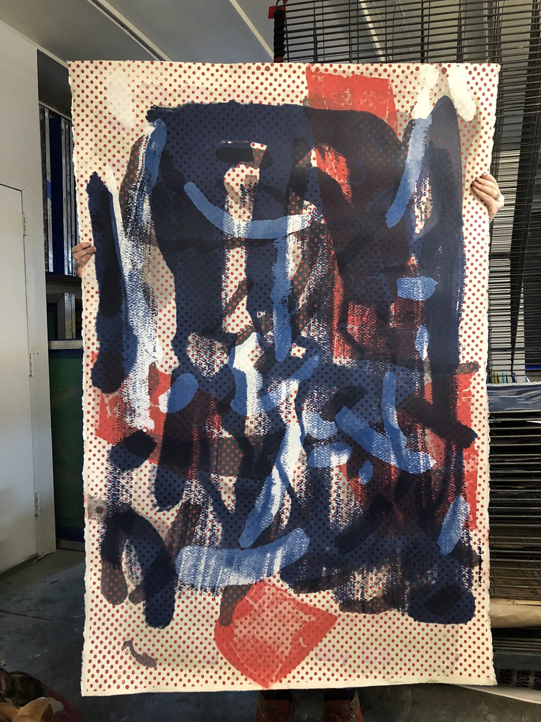 Abstract painting with bold blue and red brushstrokes over a dotted background, held up by a person in an art studio.