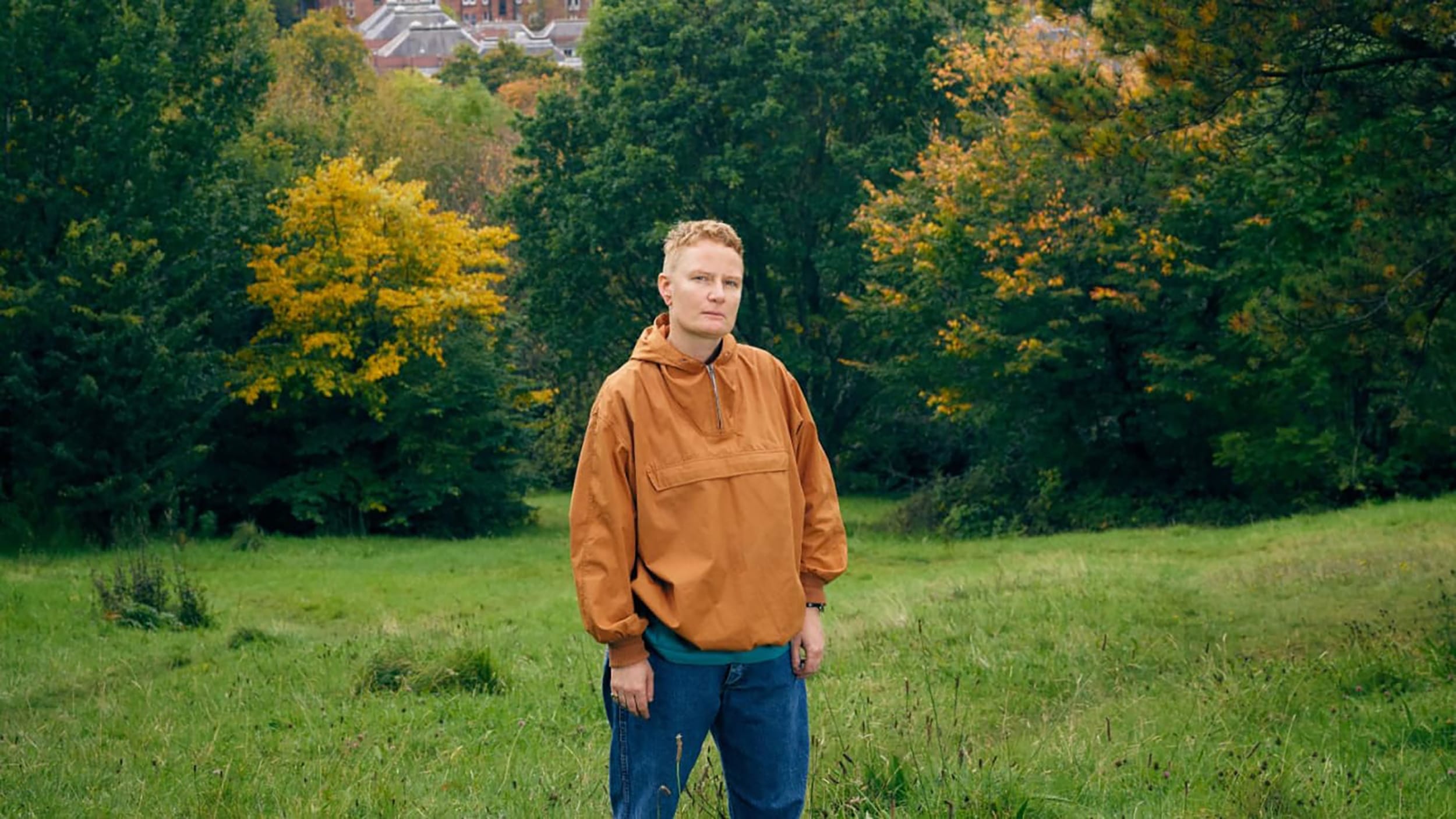 Person in a brown jacket standing in a grassy field with colorful autumn trees in the background.