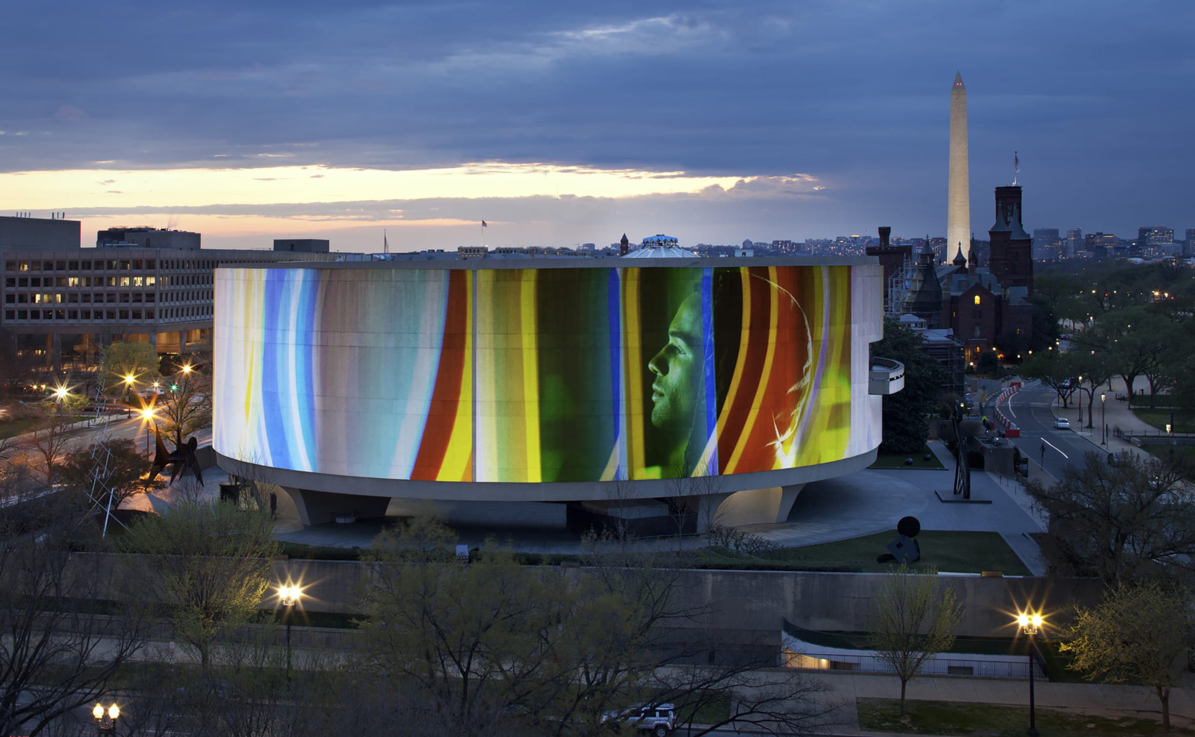 Modern museum building with digital art display at dusk, Washington Monument visible in background, surrounded by cityscape.