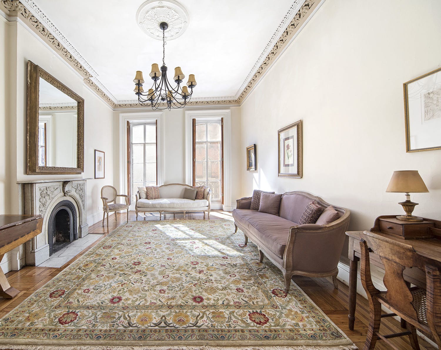 Elegant living room with vintage furniture, ornate rug, marble fireplace, and chandelier. Natural light from tall windows.