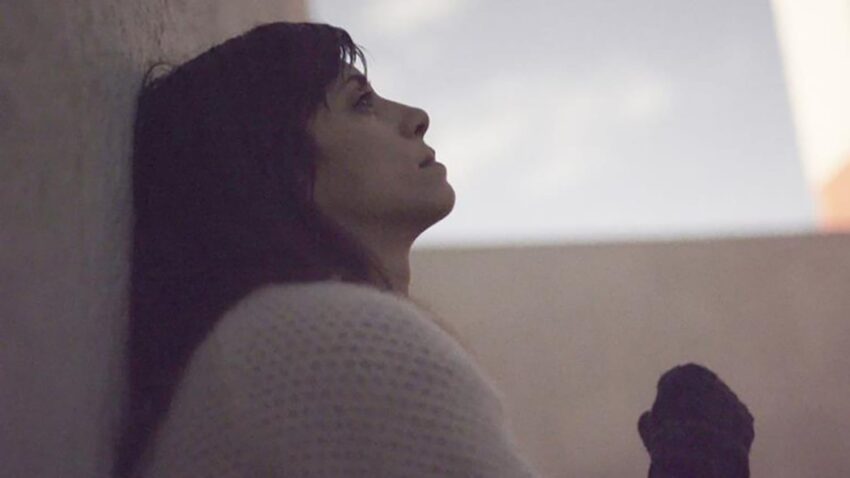 Woman in a cozy sweater leans against a wall, looking thoughtfully into the distance.