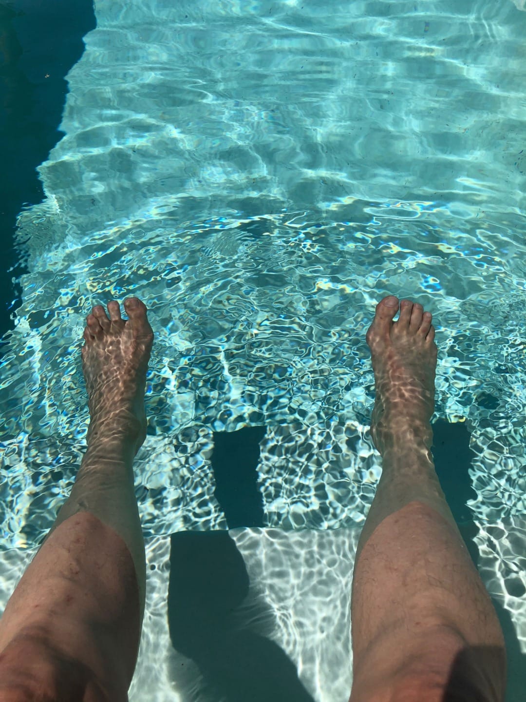 Feet submerged in a clear swimming pool, casting rippling shadows on a sunny day.