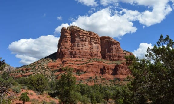 Red rock formation against a blue sky with clouds and surrounding greenery in Sedona, Arizona.