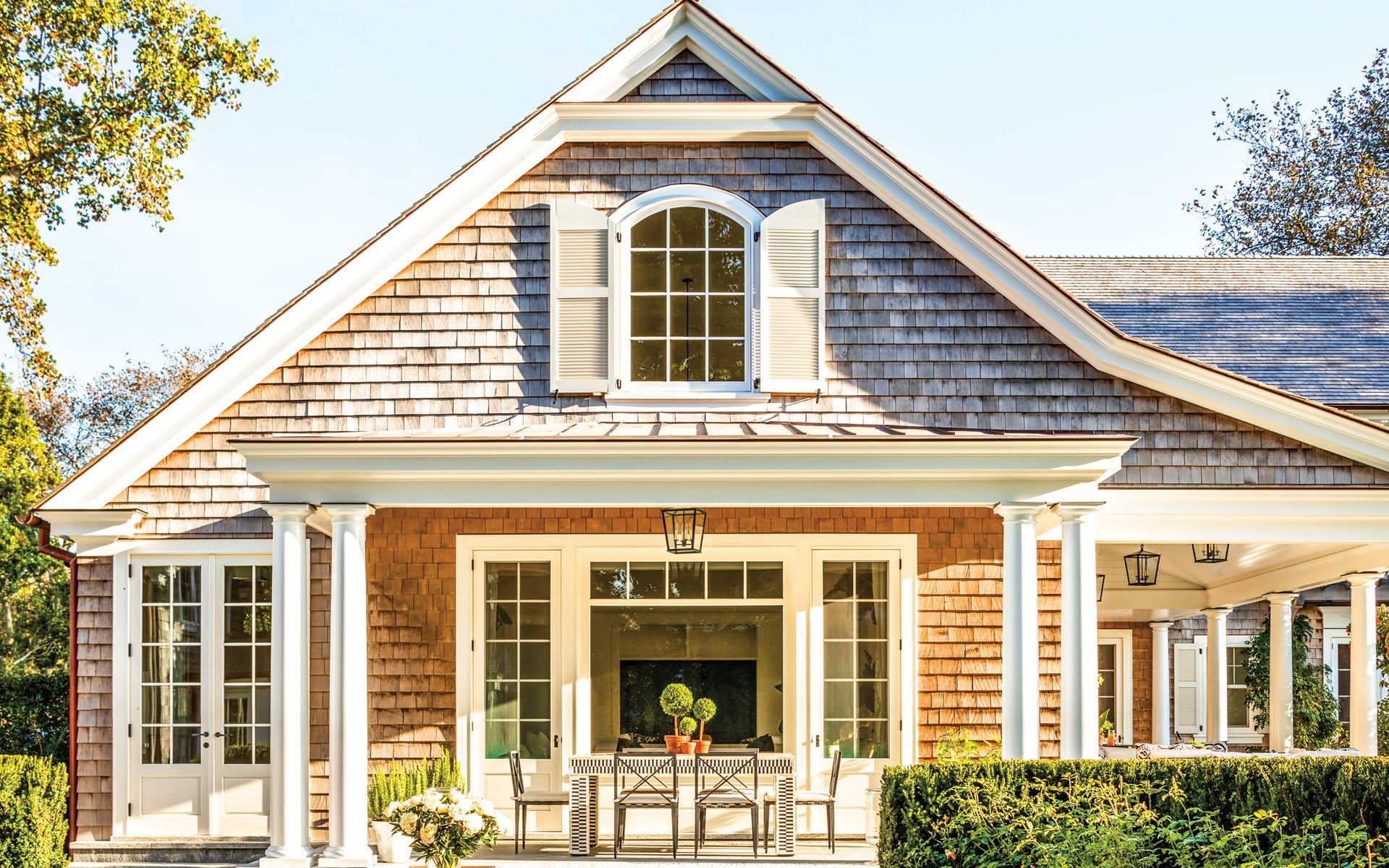 Cottage with shingle siding, large windows, and a porch with a dining table, surrounded by greenery.