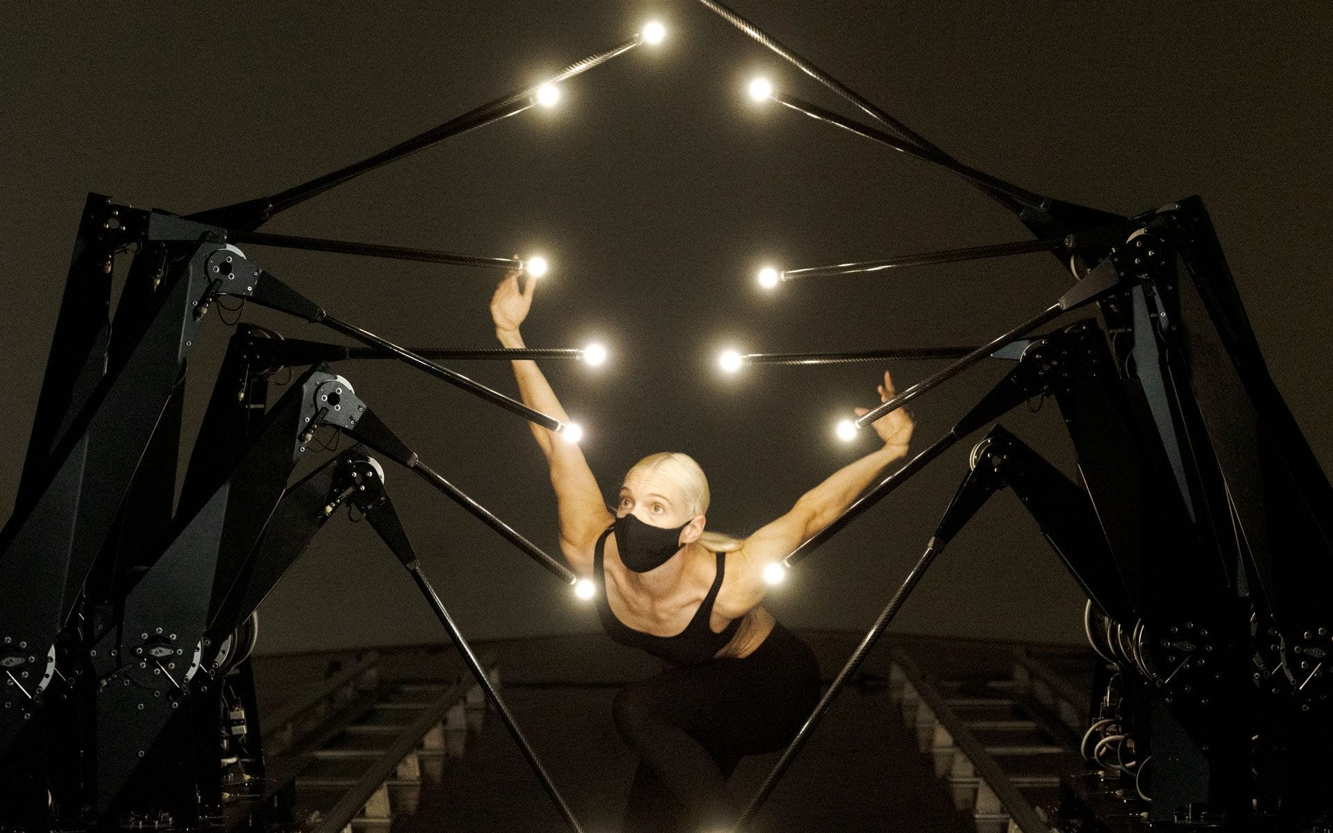 Person with short blond hair and mask performing inside a large spider-like light structure on a dimly lit stage.