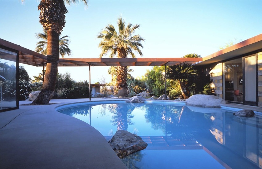 Modern house with a glass-walled pool area, surrounded by palm trees and desert landscape at sunset.