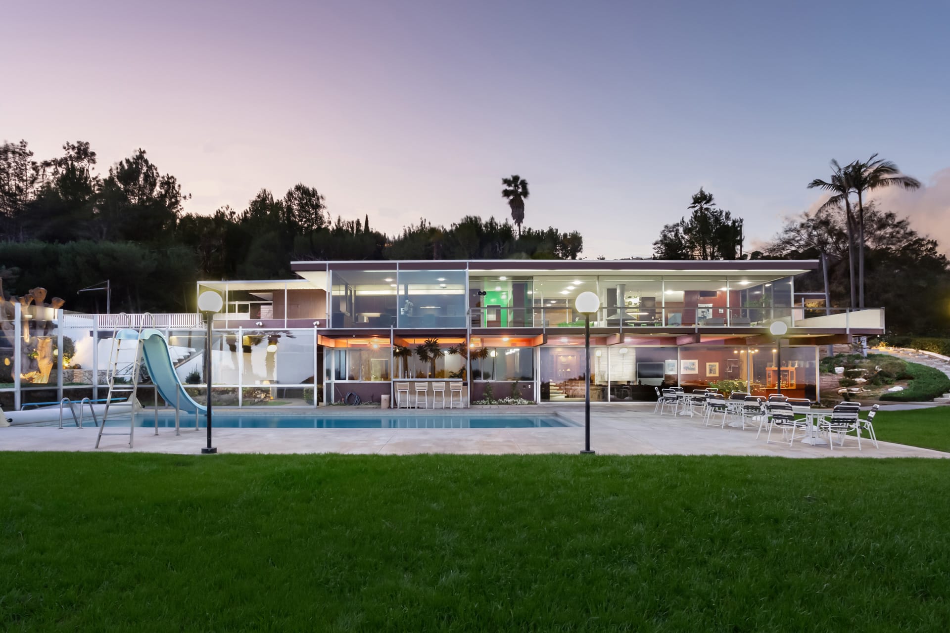 Modern glass-fronted house with large windows, outdoor pool, and patio furniture set in a lush green garden at sunset.