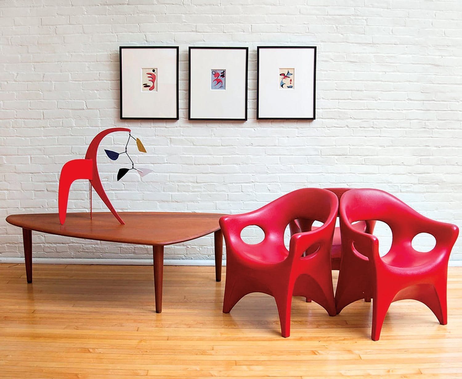 Modern room with two red chairs, a wooden table with a sculpture, and three framed artworks on a white brick wall.