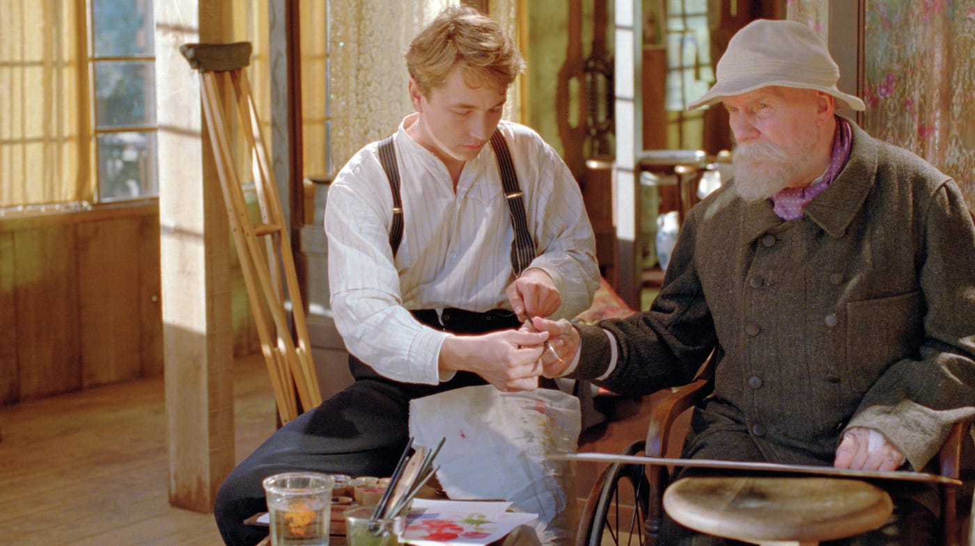Young man assists an elderly man with painting inside a sunlit room filled with art supplies and natural light.