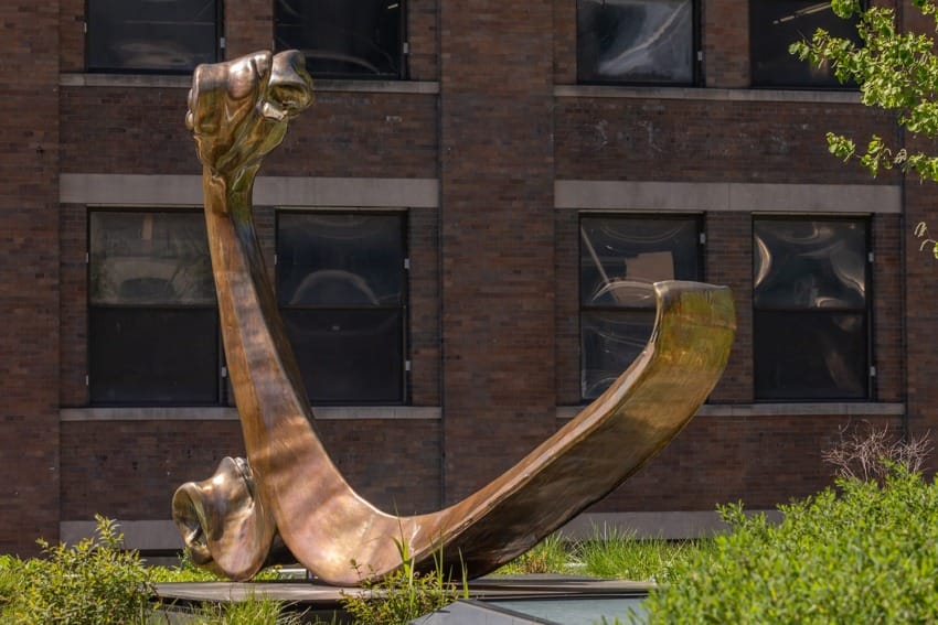 Bronze abstract sculpture of a large curved shape in a garden setting with a brick building in the background.