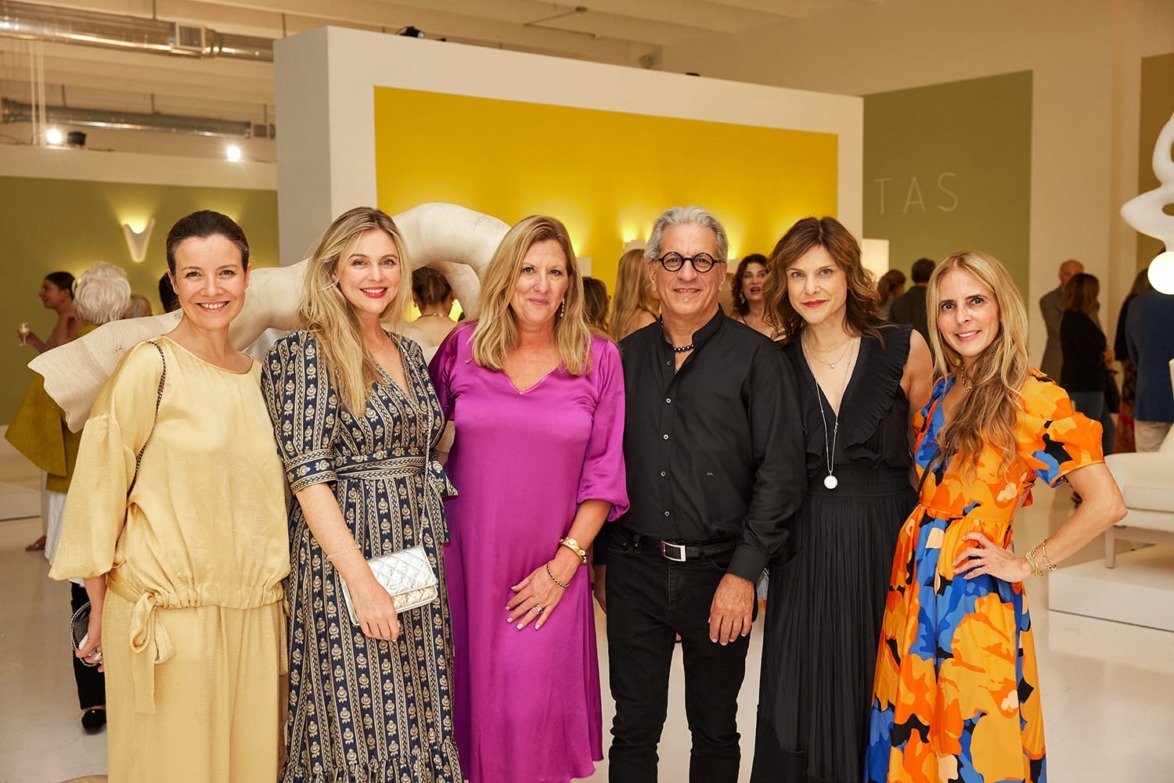 Group of six people smiling in a brightly lit art gallery setting with colorful clothing and modern decor.