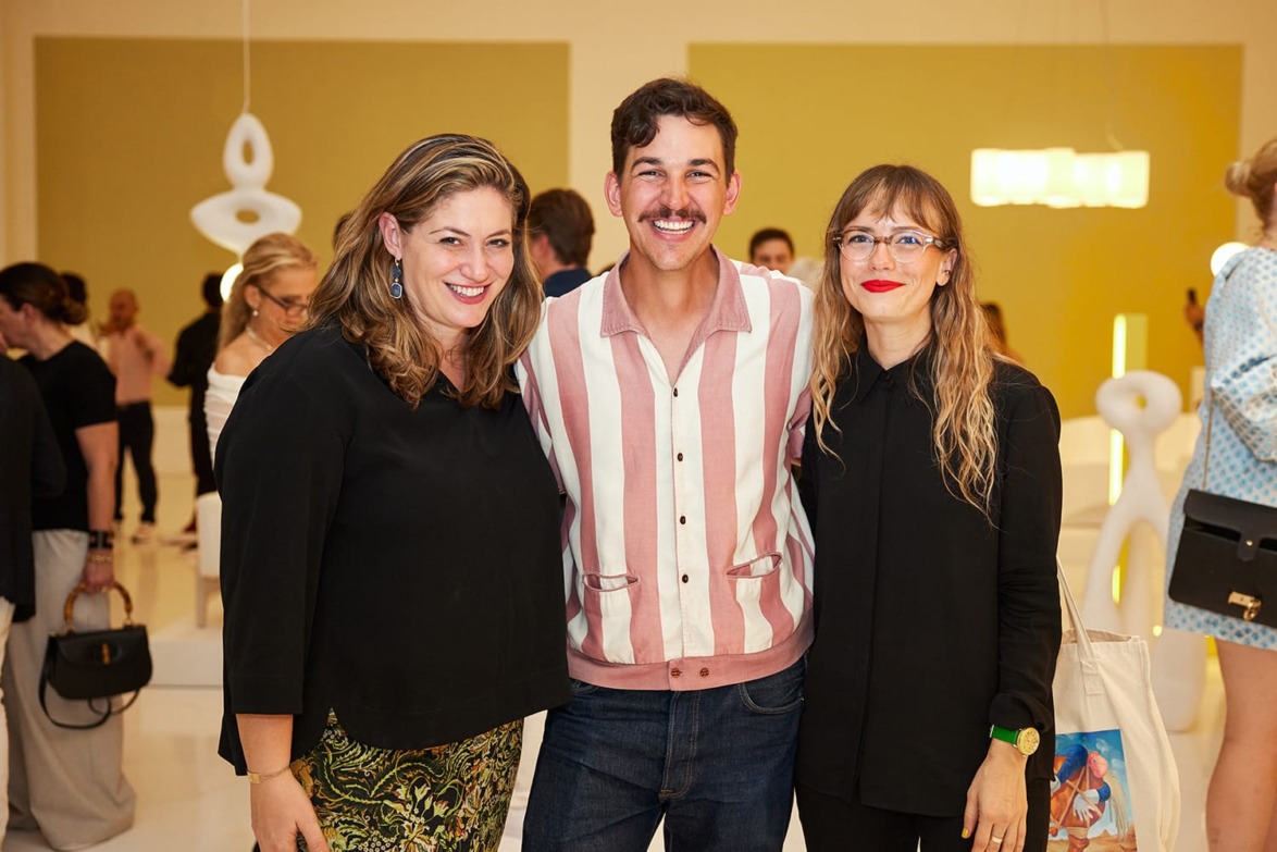 Three people smiling at an art gallery event with abstract sculptures displayed in the background.