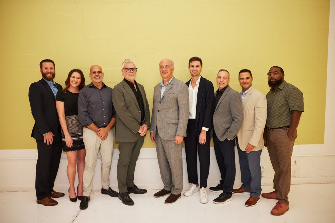 A group of nine people standing in a line against a light green wall, all smiling and dressed in semi-formal attire.