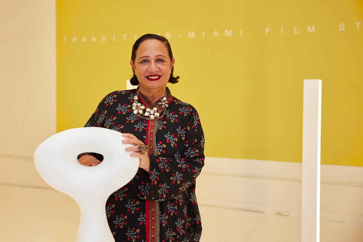 Person in floral outfit standing with modern white sculpture against a yellow backdrop with "Transitus Miami Film Studio" text.