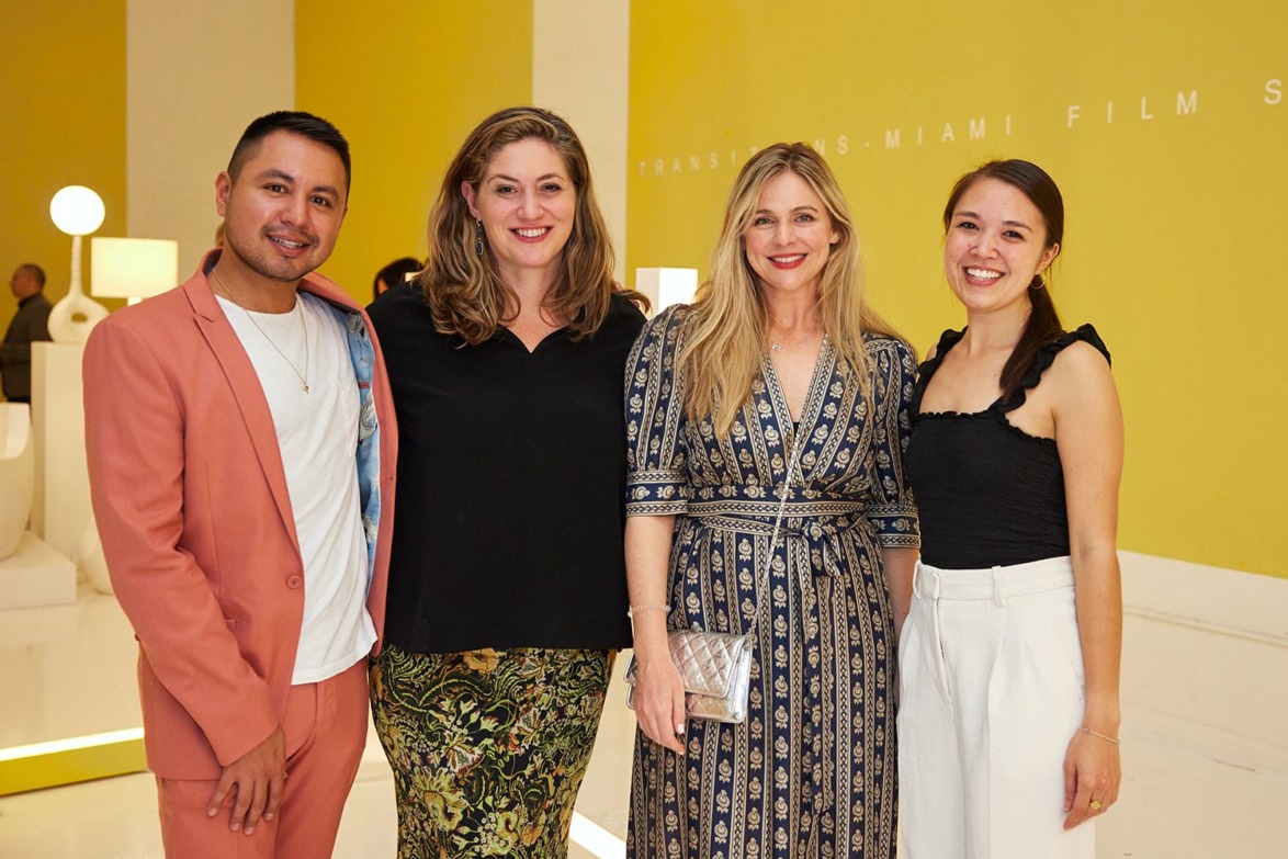 Four people smiling in front of a yellow wall at an event.