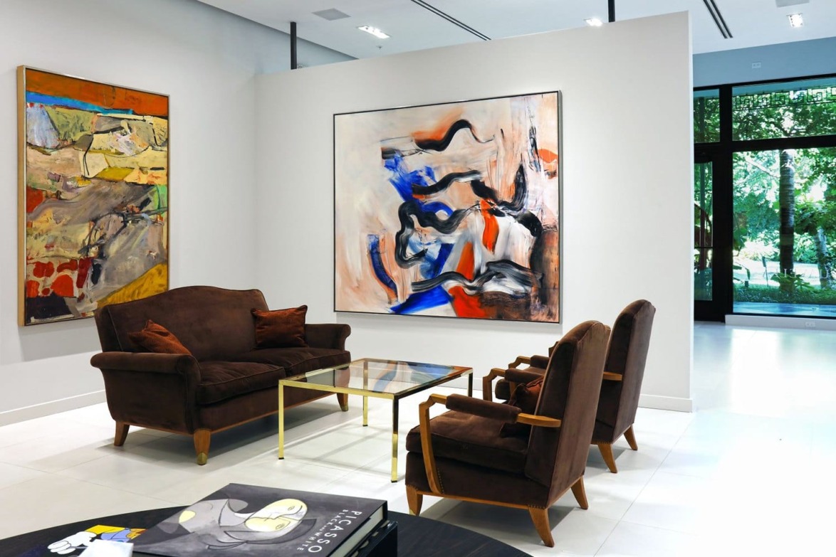 Gallery room with abstract paintings on white walls, brown sofa set, and a glass coffee table.