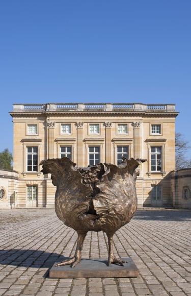 Bronze abstract chicken sculpture on cobblestone square with historic building in the background under a clear blue sky.