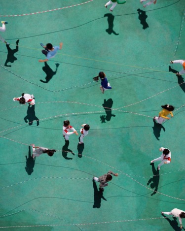 Aerial view of children skipping rope on a green playground, creating shadows of different shapes and movements.