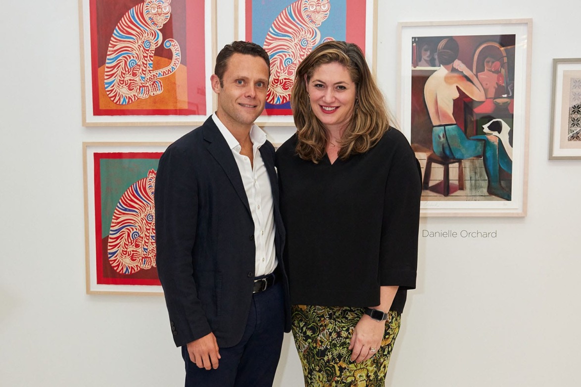 Two people pose in front of colorful framed artwork at a gallery exhibition.