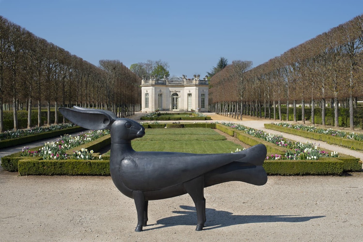 Large black bird sculpture in a formal garden with manicured trees and a historic building in the background.