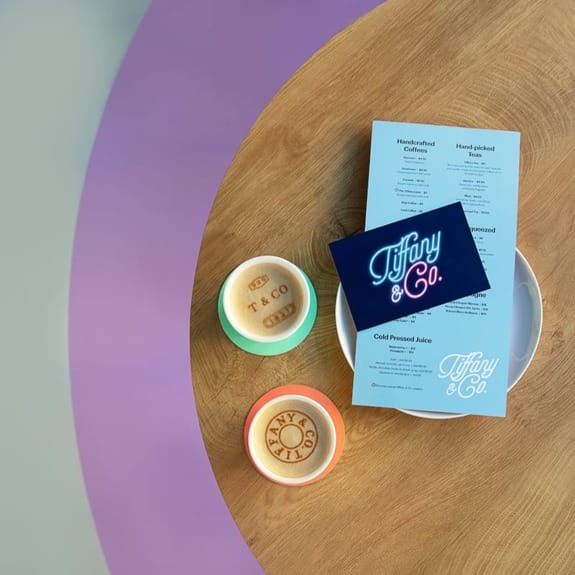 Menu with Tiffany & Co. branding on a plate, accompanied by two latte cups on a wooden table with a purple accent.