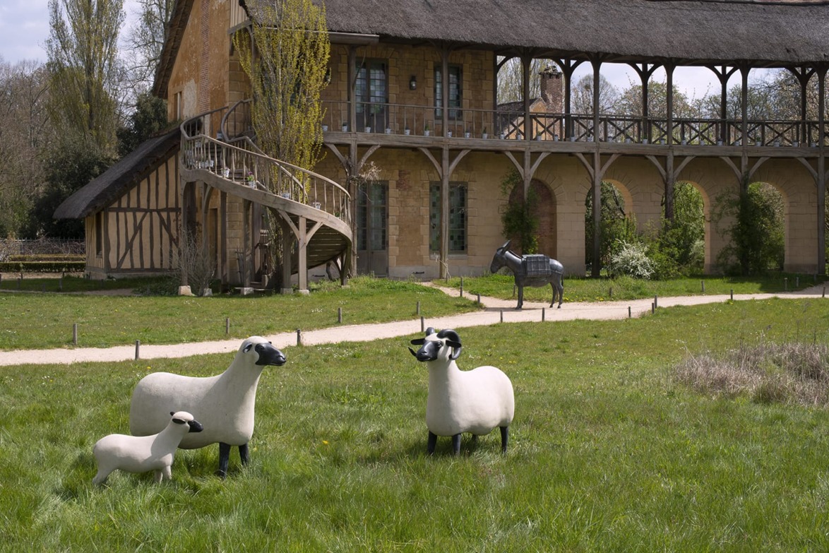 Three sheep sculptures and a donkey sculpture in front of a rustic building with arched walkways and a thatched roof.