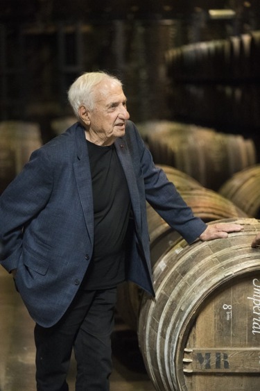 Man in a blue blazer stands next to wooden barrels in a dimly lit room.