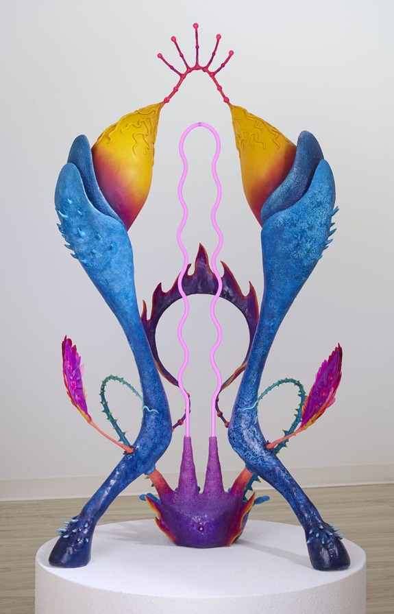 Colorful abstract sculpture with organic shapes and vibrant hues on a white pedestal in a gallery setting.