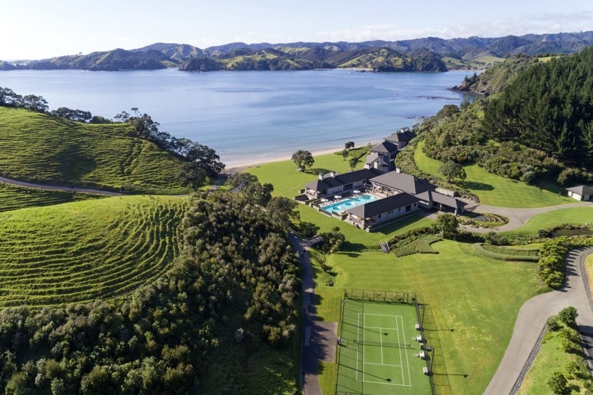Aerial view of a coastal luxury resort with tennis courts, swimming pool, and lush green hills by the ocean.