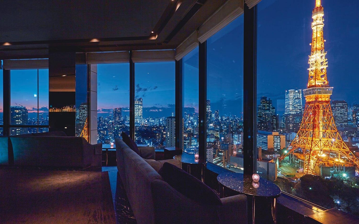 View of Tokyo cityscape at night with illuminated Tokyo Tower seen through large windows from a luxurious, dimly lit room.