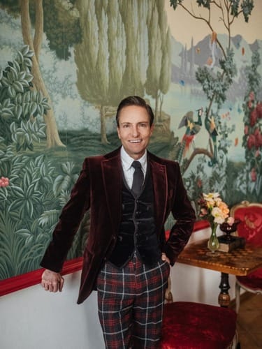 Person in a velvet jacket and plaid pants standing in a room with a nature-themed wall mural and elegant furniture.
