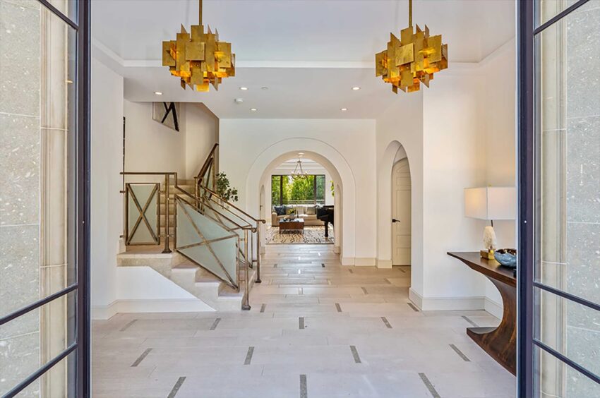 Spacious modern foyer with elegant staircase, arched doorway, and golden chandeliers leading to a bright living area.