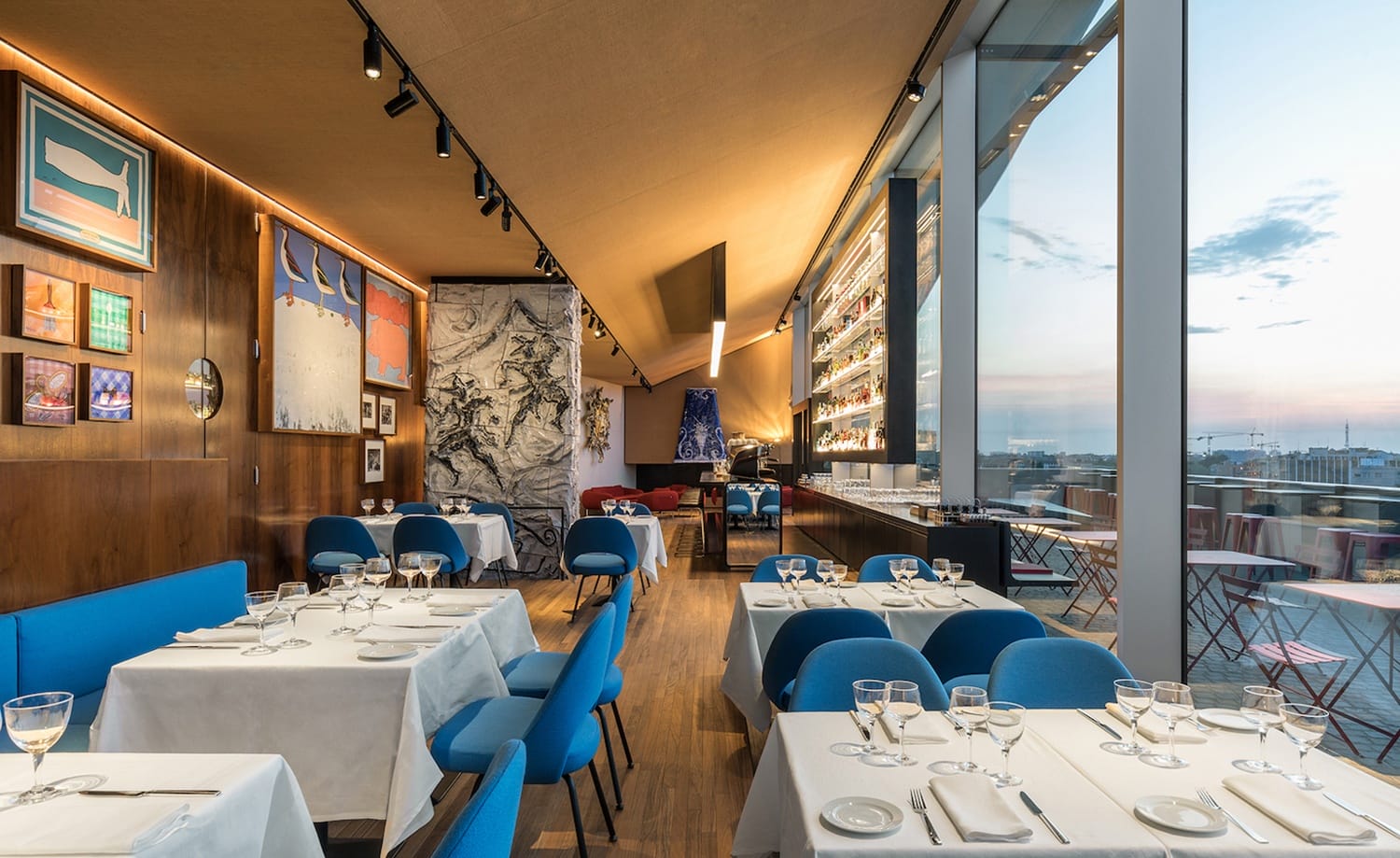 Modern restaurant interior with blue chairs, white tablecloths, and wall art, featuring large windows with a cityscape view.