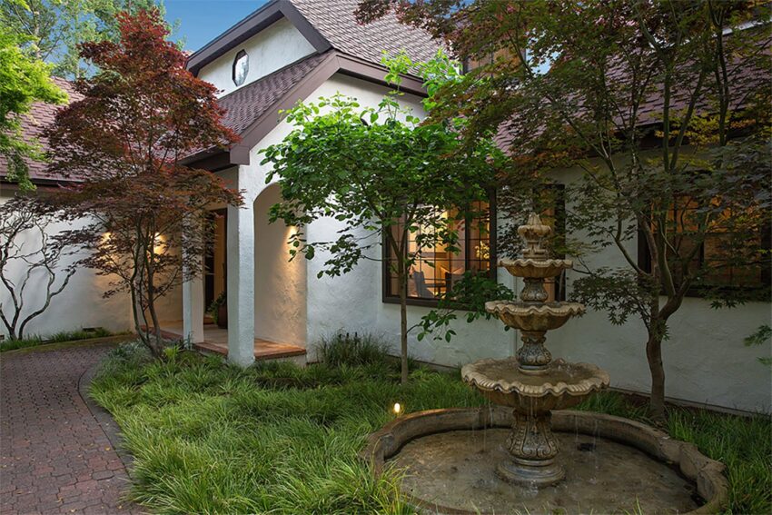 Charming cottage-style home with trees, a brick path, and a tiered fountain in the front yard.