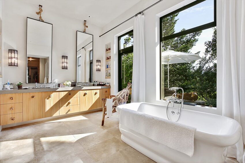 Luxurious bathroom with freestanding tub, double sinks, large mirrors, and natural light from floor-to-ceiling windows.