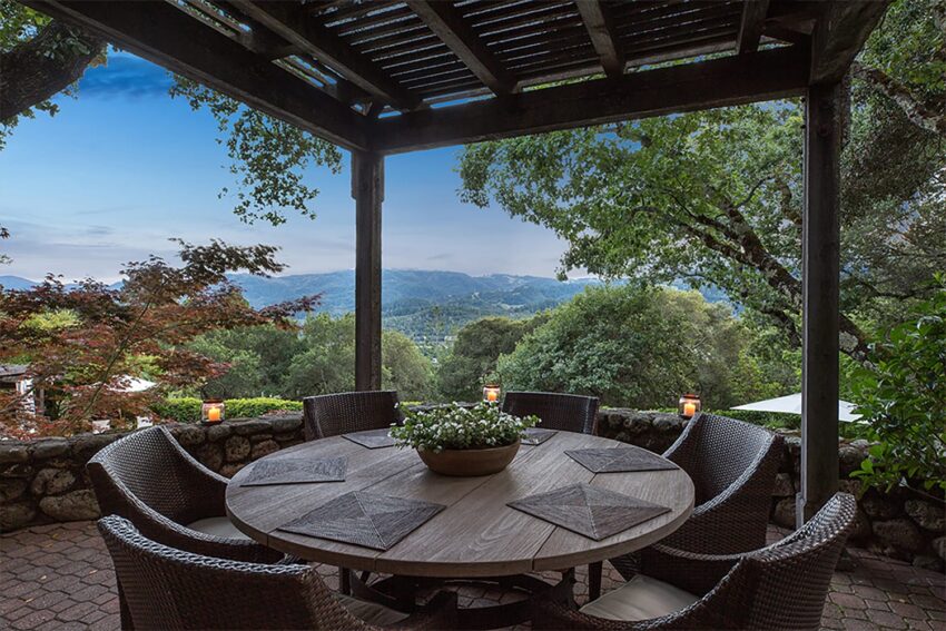 Outdoor patio with round table and wicker chairs overlooking a scenic view of mountains and lush greenery under a pergola.