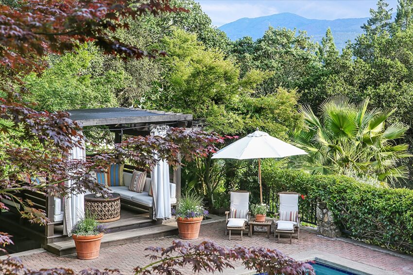 Outdoor patio with shaded seating area, two lounge chairs, umbrella, potted plants, and lush greenery in the background.