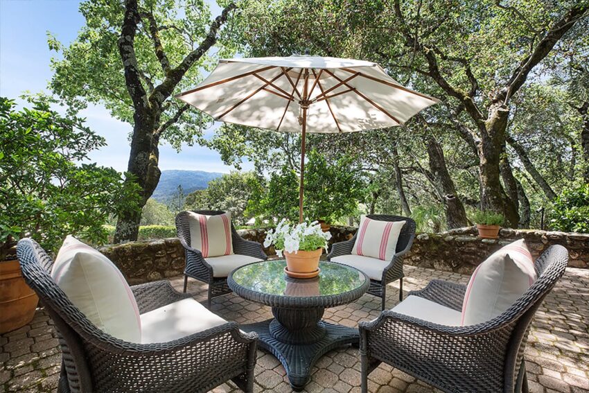 Patio set with wicker chairs and umbrella under trees, overlooking a scenic view.